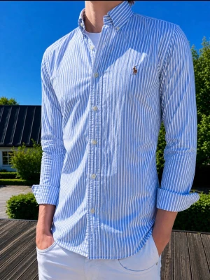 Blårandig skjorta från Ralph Lauren - (MODELLEN ÄR 186CM OCH VÄGER 75KG) OcKlassisk blårandig skjorta från Ralph Lauren med button-down krage och broderad logga på bröstet. Skjortan har långa ärmar, vita knappar och är tillverkad i bomull. Perfekt för en stilren och avslappnad look.