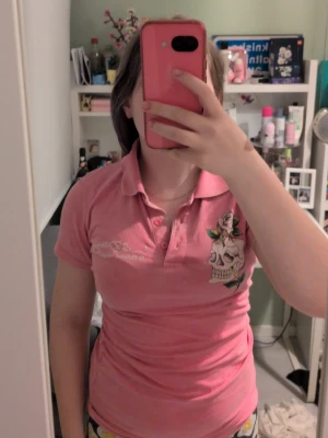 Rosa t-shirt med döskalle och blommor - Säljer en rosa t-shirt med krage och knappar framtill. På bröstet finns ett tryck med döskalle och rosor, och på ryggen ett större blommotiv. T-shirten har korta ärmar och en slim passform. Perfekt för dig som gillar unika detaljer och färg. Den är vintage. Står ingen storlek men passar som S. Vissa stenar är borta. Fråga gärna om mer info eller lägg ett bud☺️