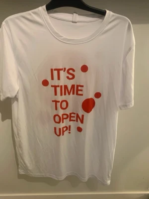 Vit t-shirt med röd text - Vit t-shirt med röd text och prickar på framsidan där det står 'IT'S TIME TO OPEN UP!'. Klassisk rund halsringning och korta ärmar. Perfekt för en avslappnad och trendig stil.