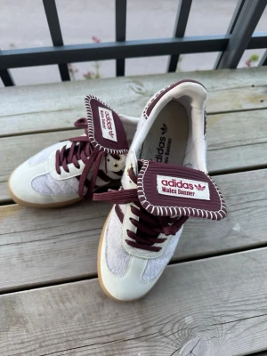 Adidas Wales Bonner sneakers vita/vinröda - Unika Adidas Wales Bonner sneakers i vitt och vinrött med grova kontrastsömmar och bred plös. Skorna har ovandel i mocka och textil, vinröda snören och detaljer samt klassisk gummisula. Perfekta för dig som vill sticka ut med en retroinspirerad look.