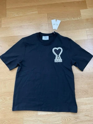 Svart t-shirt från Ami Paris - Svart t-shirt från Ami Paris med ett stort vitt hjärta och bokstaven A broderat på bröstet. Klassisk rund halsringning och korta ärmar. Tillverkad i mjuk bomull för en skön känsla och stilren look.