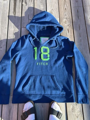 Blå hoodie med grön text från Fitch - Snygg blå hoodie med huva och känguruficka framtill. På bröstet finns ett stort grönt broderat 18 och texten FITCH. Perfekt för en avslappnad och sportig stil. Mjuk bomullskänsla och dragsko i huvan.siter mer som M/S