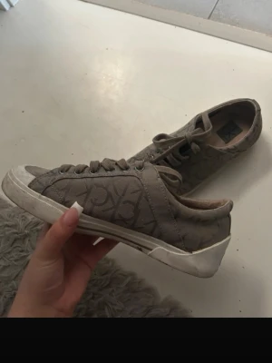 Calvin Klein sneakers  - Säljer ett par beige sneakers med tydligt logomönster och vita sulor. Skorna har klassisk låg design, snörning framtill och är tillverkade i textilmaterial. Perfekta för en avslappnad och trendig stil.