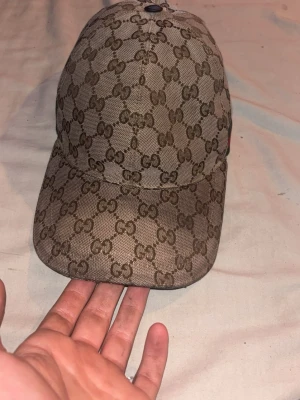 Gucci beige keps med GG-mönster - Snygg beige keps från Gucci med det klassiska GG-monogrammet över hela kepsen. På sidan finns en grön och röd rand som ger extra lyxig känsla. Justerbar rem baktill för perfekt passform. Tillverkad i canvasmaterial.