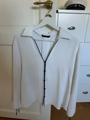 Vit skjorta med snörning framtill - Stilren vit skjorta med svart snörning längs öppningen framtill och vid ärmsluten. Skjortan har klassisk krage och lång ärm, och är tillverkad i ett mjukt, lätt material som ger en avslappnad men trendig look.