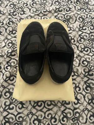 Axel arigato skor - Säljer ett par svarta slip-on sneakers med bred resår över foten och tjock vit platåsula. Skorna har en enkel och clean design, perfekta för en avslappnad streetstyle. Materialet ser ut att vara mocka eller liknande tyg. Lätta att ta på och av. Det finns ett litet hål som går in i skon på en av skorna men det gör ingen skillnad, det kan komma in nån sten ibland. Går säkert att fixa hos en skomakare. Pris går att diskuteras och fler bilder kan skickas, bara att höra av sig.