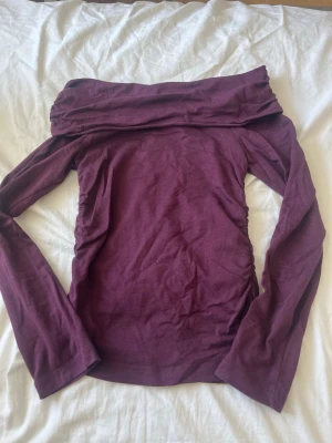 Mörklila offshoulder topp - Snygg vinröd/mörk lila offshoulder topp med långa ärmar. Toppen har en bred krage som vilar över axlarna och är tillverkad i ett mjukt bomullsmaterial. Perfekt för dig som vill ha en stilren och trendig look.