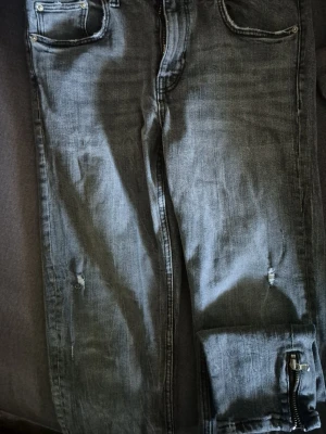 ZARA Grå slitna jeans med dragkedja - Säljer ett par grå jeans från ZARA med slitningar på knäna och dragkedjedetalj vid benslutet. Jeansen har klassisk femficksmodell och en tvättad look som ger en avslappnad vibe. Perfekta för dig som gillar streetstyle och detaljer.