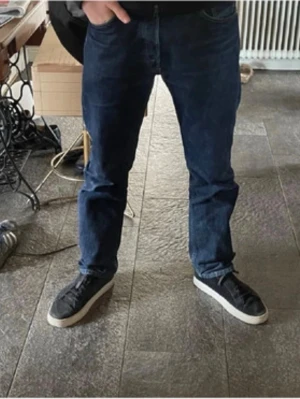 Levi's 501 mörkblå jeans herr - Klassiska Levi's 501 jeans i mörkblå denim. Rak passform med normal midja och knappgylf. Tillverkade i 100% bomull för en autentisk känsla. Perfekta för dig som gillar tidlös stil och vill ha ett par ikoniska jeans i garderoben.