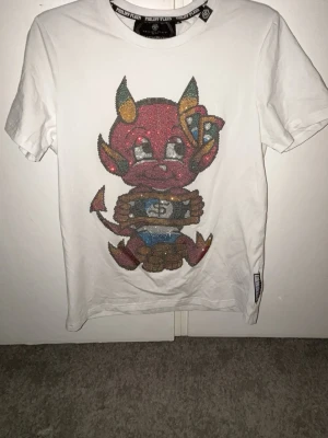 Vit Philipp Plein t-shirt med djävulstryck - Säljer en vit t-shirt från Philipp Plein med ett stort glittrigt tryck av en tecknad djävul på framsidan. T-shirten har korta ärmar och rund hals. Perfekt för dig som gillar statement-plagg och coola detaljer.