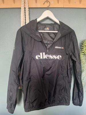 Svart vindjacka från Ellesse - Snygg svart vindjacka från Ellesse med stor logga framtill och mindre logga på bröstet. Jackan har huva med dragsko, halv dragkedja och elastiska muddar. Tillverkad i lätt och tåligt material, perfekt för blåsiga dagar.