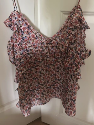 Blommig volangtop från Zara - Söt blommig topp från Zara med tunna axelband och volangdetaljer längs sidorna. Toppen har ett luftigt material och ett färgglatt blommönster i rosa, vitt och blått. Perfekt för varma dagar och ger en romantisk vibe till din outfit.