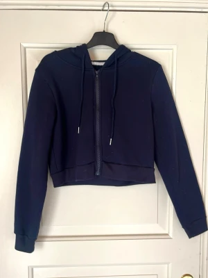 Mörkblå croppad hoodie med dragkedja - En stilren mörkblå croppad hoodie med dragkedja framtill och justerbar huva med snören. Hoodien har långa ärmar och ribbade muddar vid ärmslut och nederkant. Perfekt för en avslappnad och trendig look. Materialet känns mjukt och bekvämt.