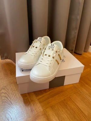 Vita Valentino sneakers med guld nitar - Snygga vita sneakers från Valentino med guldfärgade nitar på ovandelen och sidan. Skorna har en klassisk låg profil, rund tå och vita snören. Tillverkade i slätt läder för en clean och exklusiv look. Perfekta för dig som vill sticka ut med detaljer. Allt medföljer, fler storlekar finns