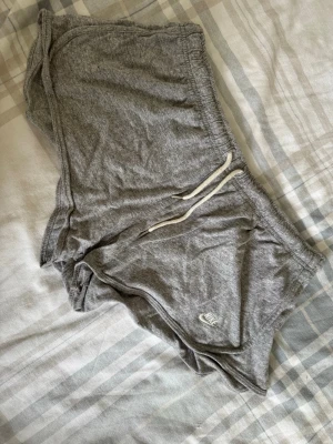 Grå mjukisshorts från Nike XL - Grå mjukisshorts från Nike med vit snörning i midjan och diskret logga på benet. Tillverkade i en mjuk bomulls- och polyestermix. Perfekta för chill eller träning, med relaxed passform och elastisk midja.