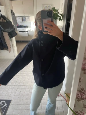 Svart rock med hög krage och knappar - Stilren svart rock med hög krage och stora knappar framtill. Rocken har en rak passform och vida ärmar, vilket ger en modern och avslappnad look. Perfekt för dig som gillar minimalistisk stil och vill ha något som sticker ut lite extra.