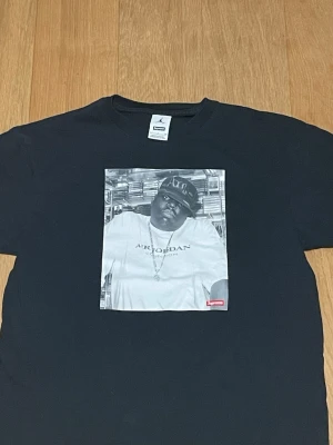 Supreme X Air Jordan Biggie svart - Supreme t-shirt i svart med stort grafiskt tryck på framsidan som visar en person i Air Jordan t-shirt och keps. Klassisk rund hals och korta ärmar. Materialet är bomull och Supreme-loggan syns tydligt på trycket.
