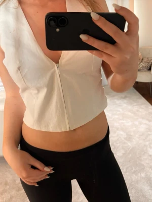 Vit croppad korsett med snörning - Trendig vit croppad korsett med dragkedja framtill och snygg snörning i sidorna med bruna detaljer. Djup v-ringning och ärmlös design, perfekt för att skapa en edgy look. Passar dig som gillar stilrena och moderna toppar.