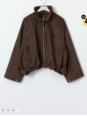 Brun mockajacka med hög krage - Snygg brun jacka i mockaimitation med hög krage och dragkedja framtill. Jackan har en loose passform, justerbar midja med spännen och två fickor fram. Perfekt för dig som gillar en trendig och avslappnad stil. 