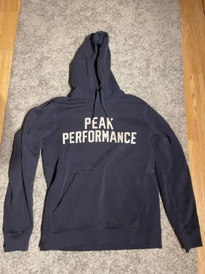 Mörkblå Peak Performance hoodie - Mörkblå hoodie från Peak Performance med stor vit logga på bröstet. Klassisk modell med huva och känguruficka framtill. Mjuk bomullsblandning och ribbade muddar vid ärmslut och nederkant. Perfekt för en avslappnad och sportig stil.