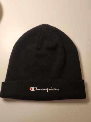 Svart mössa från Champion - Snygg svart mössa från Champion med broderad logga framtill i vitt och rött. Klassisk modell med uppvikt kant, perfekt för att hålla värmen och ge en sportig vibe. Passar till de flesta outfits och är enkel att matcha.