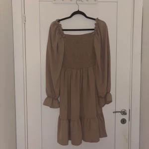 Beige långärmad volangklänning - Söt beige klänning med långa puffärmar och volangdetaljer nertill. Smockad överdel och rundad halsringning ger en trendig look. Perfekt för dig som gillar en romantisk och avslappnad stil.