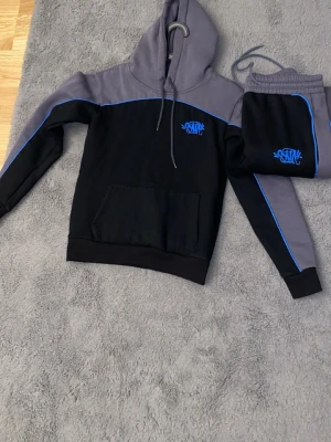 Svart och grått loungewear-set OSTAIC - Snyggt loungewear-set från OSTAIC med hoodie och matchande byxor i svart och grått med blå detaljer. Hoodien har magficka och dragsko, byxorna har resår i midjan. Perfekt för chill dagar hemma.