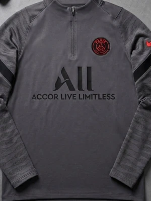 Grå långärmad PSG träningströja - Säljer en grå långärmad träningströja från Nike med PSG-logga på bröstet och Accor Live Limitless-tryck. Tröjan har halv dragkedja, svarta detaljer på ärmarna och diskret mönster. Perfekt för fotbollsträning eller sport.(SLIM FIT)