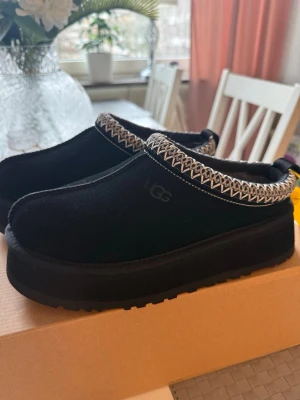 Svarta UGG tofflor med mönstrad kant - Säljer ett par svarta UGG tofflor i mocka med tjock sula och snyggt mönstrad kant i vitt och grått. Perfekta för att hålla fötterna varma hemma eller ute. Klassisk slip-on modell med UGG-logga på sidan och mjuk insida.