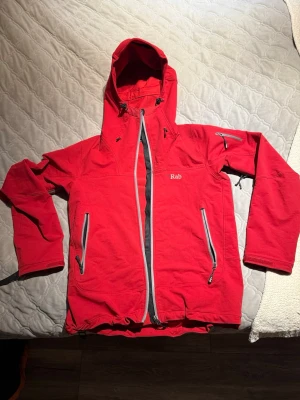 Röd softshelljacka från Rab med huva - Snygg röd softshelljacka från Rab med justerbar huva, dragkedjor i kontrastfärg och flera praktiska fickor. Jackan har kardborre vid ärmsluten och dragsko nertill för bästa passform. Perfekt för blåsiga dagar och outdoor-äventyr.