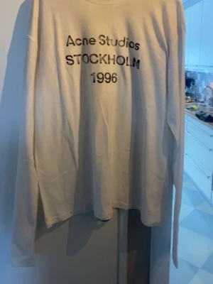 Vit långärmad topp Acne Studios - Vit långärmad topp från Acne Studios med svart tryck framtill där det står 'Acne Studios STOCKHOLM 1996'. Tunn och mjuk bomullskvalitet med rund halsringning. Perfekt för en clean och stilren look.
