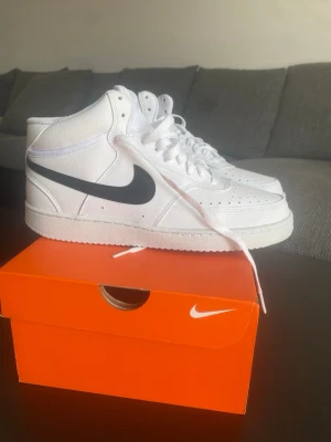 Nike vita midsneakers med svart swoosh - Fräscha vita sneakers från Nike med svart swoosh på sidan. Klassisk mid-modell med snörning och perforerade detaljer på tån. Tillverkade i slitstarkt läder och har en bekväm vadderad krage. Perfekta för dig som gillar stilrena och sportiga skor.