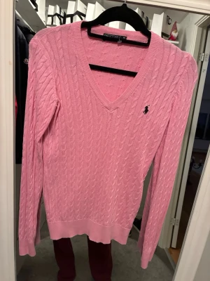 Rosa kabelstickad tröja Polo Ralph Lauren - Kabelstickad rosa tröja från Polo Ralph Lauren med V-ringning och svart broderad logga på bröstet. Tröjan har långa ärmar, ribbade muddar och är gjord i mjukt material som känns skönt mot huden. Perfekt för dig som gillar klassisk preppy stil.