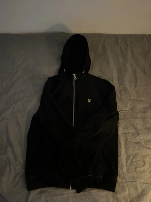 Svart vindjacka från Lyle & Scott - Snygg svart vaderad vind/regn jacka från Lyle & Scott med huva och dragkedja framtill. Jackan har en gul broderad logga på bröstet och diskreta sidofickor. Materialet är slätt och lätt, perfekt för blåsiga dagar. Enkel och stilren design som passar till det mesta.