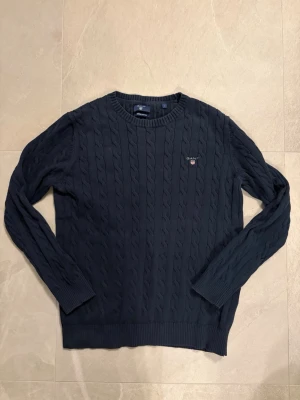 Mörkblå kabelstickad tröja från GANT - Snygg mörkblå kabelstickad tröja från GANT med rund hals och broderad logotyp på bröstet. Tröjan har ribbade muddar vid ärmslut och nederkant, och är tillverkad i mjukt material som passar perfekt för kyliga dagar.