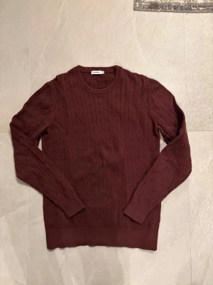 Mörkröd stickad tröja från Lindbergh - Säljer en mörkröd stickad tröja från Lindbergh med diskret kabelstickat mönster. Tröjan har rund halsringning, långa ärmar och ribbade muddar vid ärmslut och nederkant. Perfekt för dig som gillar stilrena och klassiska plagg.
