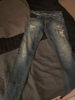 Slim fit jeans med slitningar - Snygga blå slim fit jeans i modellen Glenn med slitna detaljer och hål på låret och vid fickan. Jeansen har klassisk femficksdesign och är tillverkade i denim. Perfekta för en trendig och avslappnad stil.