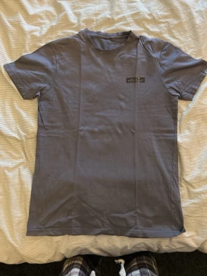 Grå/mörkblå t-shirt från Ellesse - Snygg grå t-shirt från Ellesse med klassisk rund hals och korta ärmar. T-shirten har en liten Ellesse-logga på bröstet och är gjord i mjuk bomull som känns skön mot huden. Perfekt till jeans eller shorts för en avslappnad look.