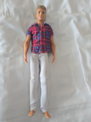 Ken-docka från Mattel 2012 - Säljer en Ken-docka från Mattel, tillverkad 2012. Dockan har blont hår och är klädd i en röd- och blå-rutig skjorta samt vita byxor. Perfekt för samlare eller som leksak. Dockan är gjord av plast och har realistiska detaljer.