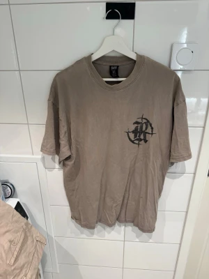 Beige t-shirt Raised by the Streets - Beige t-shirt från Raised by the Streets med svart tryck på bröstet och stor text på ryggen. Oversized passform och rund halsringning. Perfekt för dig som gillar streetwear och vill sticka ut med en unik design.