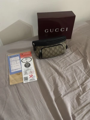 Gucci beige magväska med monogram - Säljer en snygg magväska från Gucci i beige canvas med klassiskt GG-monogram och svarta detaljer. Väskan har justerbart svart band och dragkedja upptill. Perfekt för att bära det viktigaste med stil. Originalbox och äkthetsintyg medföljer.