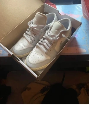 Vita Nike Air Force 1 sneakers - Säljer ett par klassiska vita Nike Air Force 1 sneakers i lågt snitt. Skorna har en stilren design med perforerad tå, vita skosnören och Nike-logga på sidan. Tillverkade i läder med gummisula för extra komfort och streetkänsla.