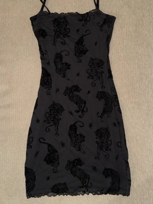 Svart kort klänning med tigerprint - Svart kort klänning från H&M med tunna axelband och ett coolt sammetsmönster av svarta tigrar. Klänningen har en tight passform och är perfekt för dig som vill sticka ut med ett djurmönster. Materialet känns mjukt och stretchigt.