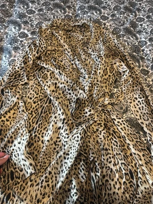 Leopardmönstrad omlottklänning - Säljer en snygg leopardmönstrad omlottklänning i beige, svart och bruna toner. Klänningen har ett tunt, flowy material och knyts i midjan för en smickrande siluett. Perfekt för dig som vill sticka ut med ett djurmönster.