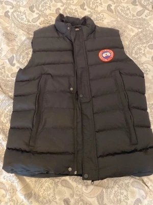 Svart dunväst från Canada Goose - Snygg svart dunväst från Canada Goose med klassisk logotyp på bröstet. Västen har hög krage, två fickor framtill och stängs med både dragkedja och knappar. Perfekt för lager-på-lager och håller dig varm under kyliga dagar.