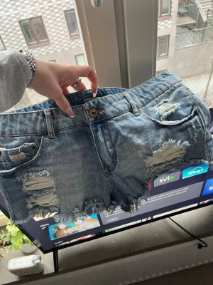 Blå slitna jeansshorts Pull&Bear - Säljer ett par blå jeansshorts från Pull&Bear med slitna detaljer och fransig kant. Klassisk femficksmodell med knapp och dragkedja. Perfekta för sommaren och ger en cool, avslappnad vibe.