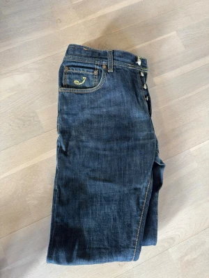 Mörkblå jeans från Jacob Cohen - Snygga mörkblå jeans från Jacob Cohen med gul kontrastsöm och klassisk femficksmodell. Jeansen har knappgylf och diskret broderad logga på myntfickan. Tillverkade i mjukt denimtyg för en bekväm känsla.