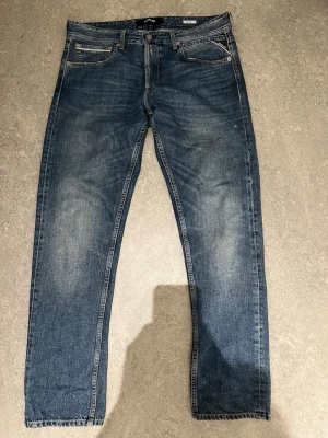 Blå raka jeans från Replay - Säljer ett par klassiska blå jeans från Replay med raka ben och fem fickor. Jeansen har snygga slitningar på framsidan och en läderpatch med logga bak i midjan. Materialet är robust denim som känns bekvämt och slitstarkt.
