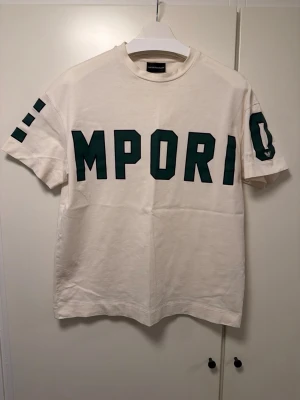Vit t-shirt från Emporio Armani - Vit t-shirt från Emporio Armani med stora gröna bokstäver över bröstet och detaljer på ärmarna. Klassisk rund hals och rak passform, perfekt för en stilren och trendig look.