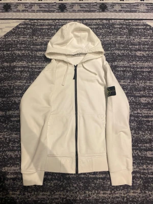 Vit Stone Island kofta - Snygg vit hoodie från Stone Island med dragkedja framtill och klassisk logotyp-patch på ärmen. Tröjan har huva med dragsko och två fickor framtill. Perfekt för en clean och stilren look.